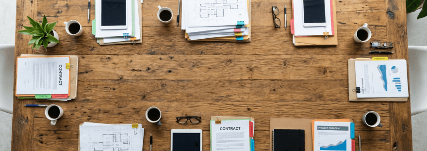 Overhead view of a wooden conference table organized with architectural plans, tablets, and coffee.