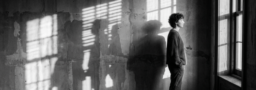 Person standing with hands in pockets looking out a window with light and shadows on a textured wall