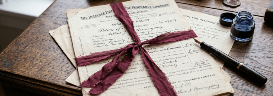 Vintage insurance documents signed by Arthur J. Hemingway tied with a red ribbon on a wooden desk next to a fountain pen and blue ink bottle